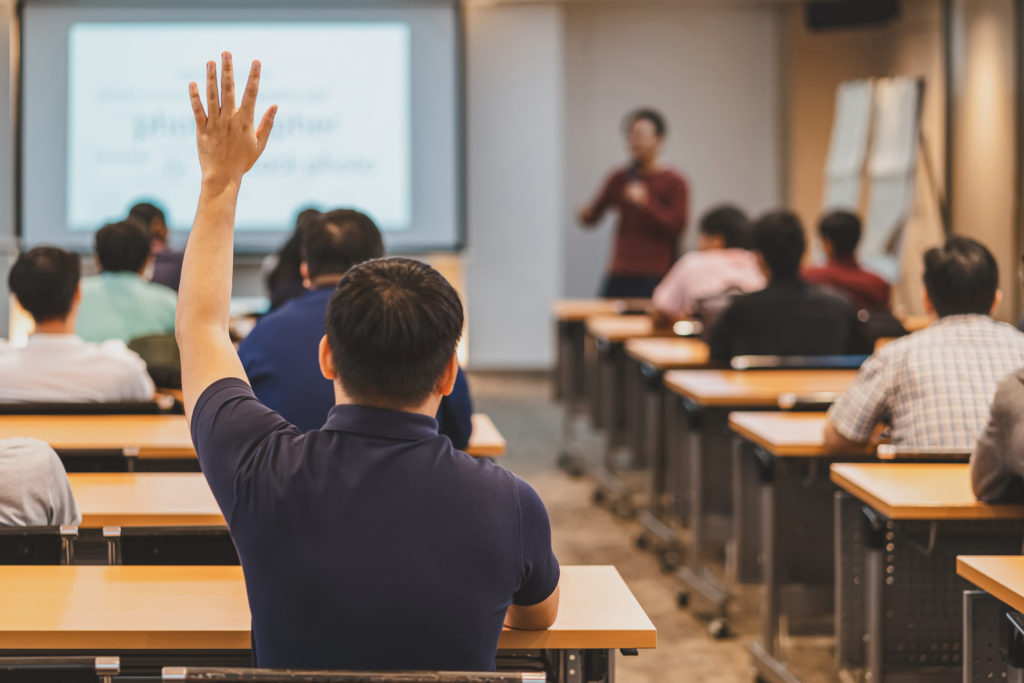 Man Raising hand in classroom of people rear view