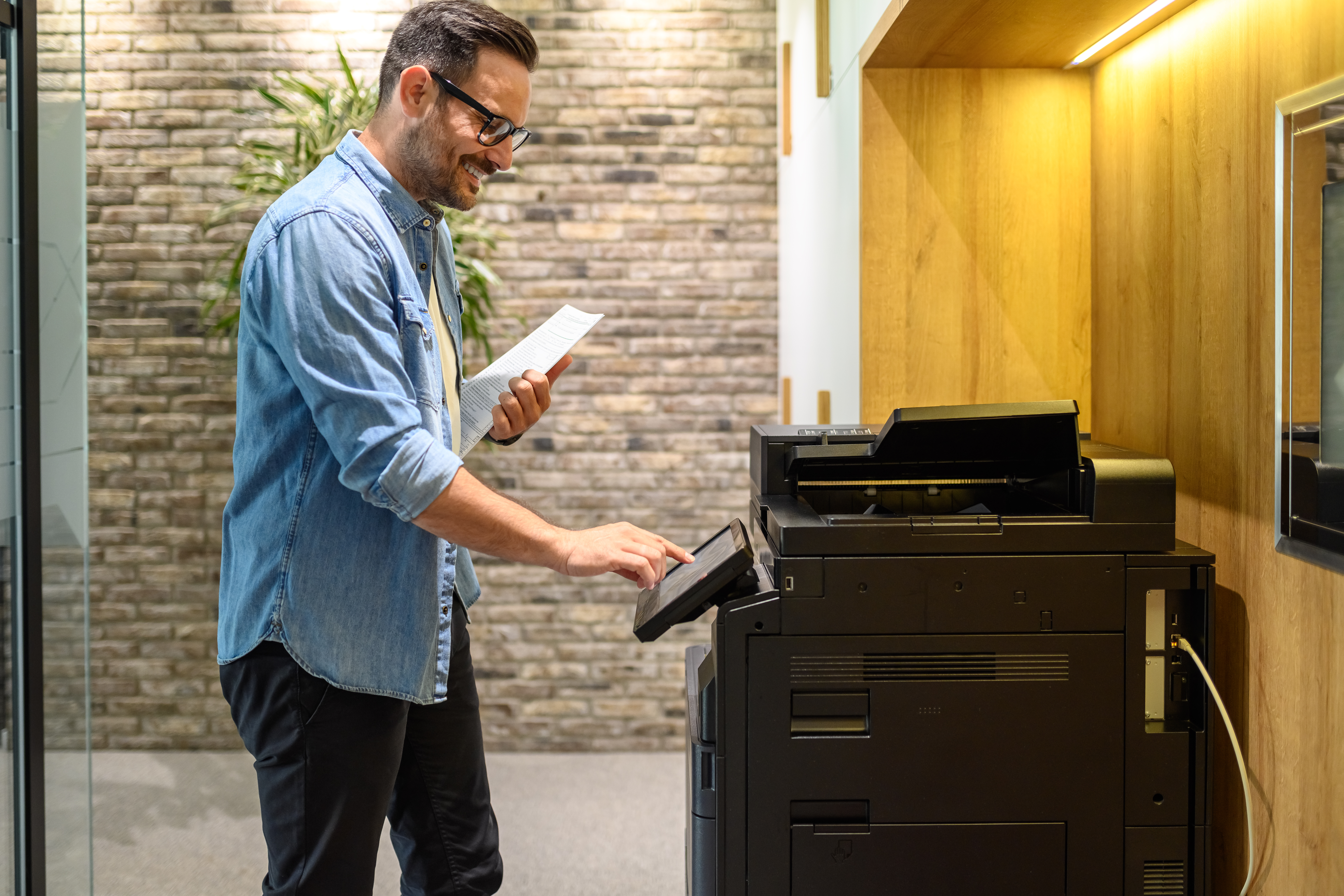Side view of smiling male professional holding documents and using photocopying machine in office